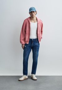 Ροζ υπερμεγέθη φούτερ, λευκή αμάνικη μπλούζα, φθαρμένα μπλε τζιν και μπεζ αθλητικά παπούτσια με καφέ λεπτομέρειες. Μπλε καπέλο με κεντητό κείμενο.