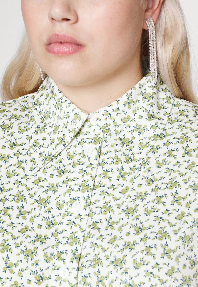 Camicia bianca con bottoni, caratterizzata da un motivo floreale in tonalità di verde e blu, con colletto classico e tessuto morbido. Orecchini luccicanti visibili.
