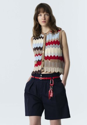 Junge Frau trägt eine ärmellose mehrfarbige Zickzack-Strickweste und marineblaue hoch taillierte Shorts mit einem roten Gürtel und Quastenaccessoire.