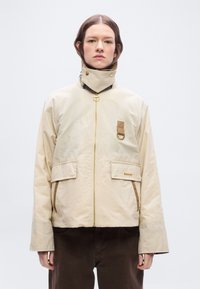 Γυναίκα που φοράει μπεζ ζακέτα με φερμουάρ και ψηλό γιακά, δύο εμπρόσθιες τσέπες με καπάκια, καφέ παντελόνι, στέκεται μπροστά σε απλό φόντο.