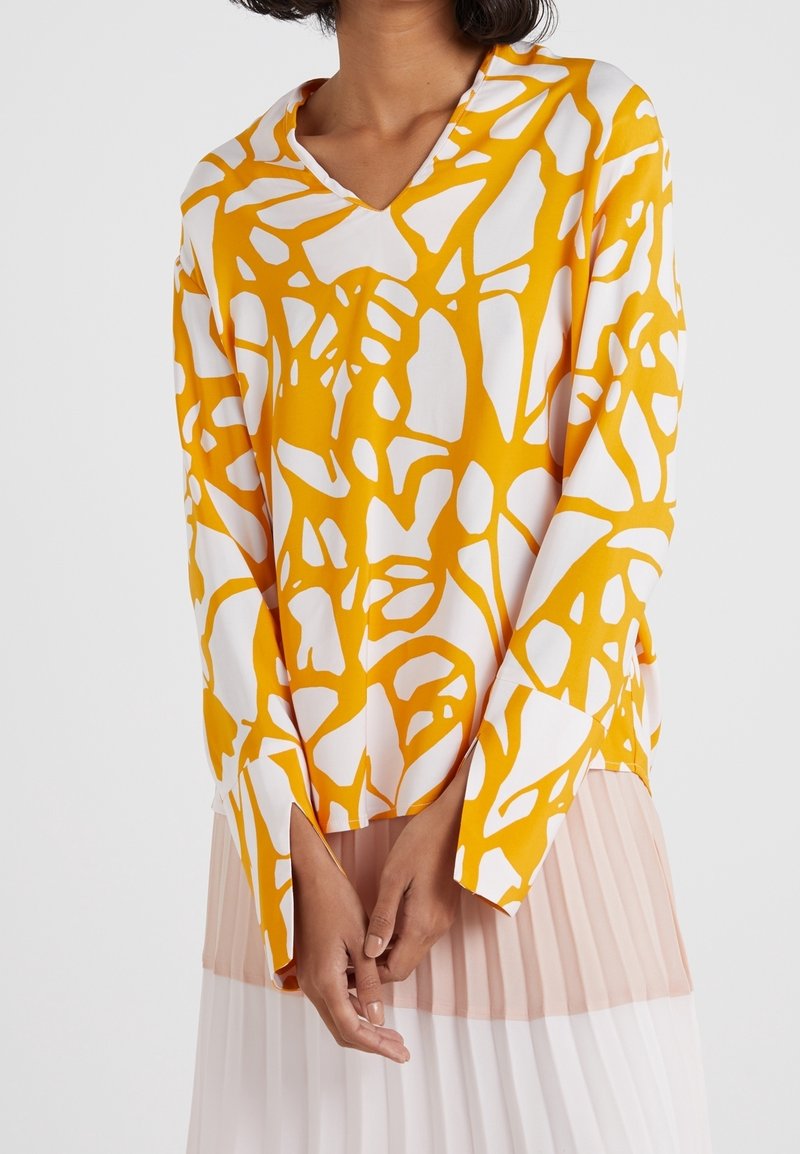 Femme portant un chemisier blanc à manches longues avec un motif jaune abstrait et une jupe plissée rose clair et blanche, mains jointes devant elle.