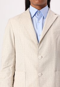 Light beige blazer with a textured pattern, two buttons, and two front pockets, paired with a light blue collared shirt underneath.