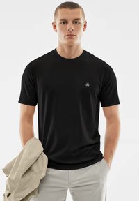 Schwarzes T-Shirt mit kurzen Ärmeln aus einem weichen Stoff, mit einem kleinen weißen Logo auf der Brust. Getragen mit hellen Hosen und einer beigen Jacke.