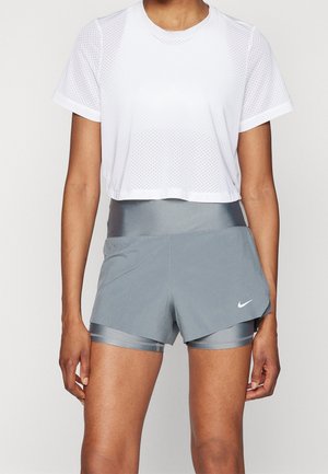 Vrouw draagt een wit kort mouwtjes mesh crop top en grijze gestapelde sportshort met hoge taille en Nike-logo op het linkerbeen.