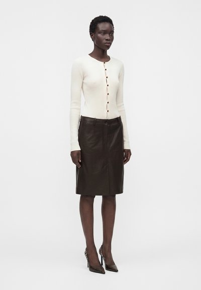 Femme portant un cardigan blanc ajusté boutonné, une jupe en cuir sombre longueur genou et des talons hauts pointus foncés, debout devant un fond uni.