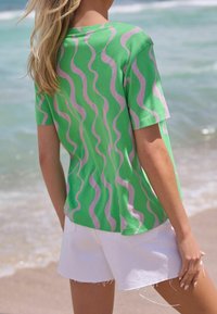 Mujer con cabello largo y rubio, vestida con una camiseta de un patrón ondulado verde y rosa y pantalones cortos blancos, de pie en una playa de arena cerca del agua.