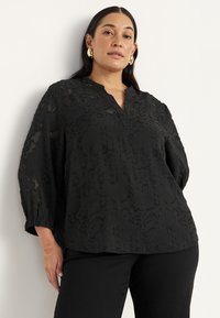 Sort blouse med et struktureret design, V-hals og trekvartærmer. Løs pasform med samlede manchetter; parret med sorte bukser.