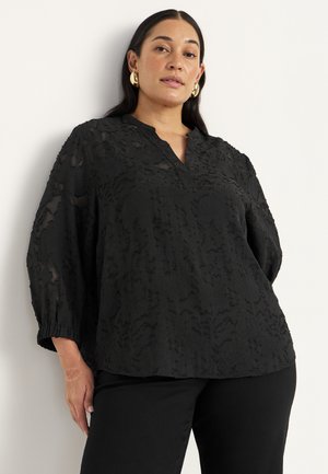 Svart blus med en texturerad design, V-ringning och trekvartsärmar. Löst snitt med rynkade manschetter; kombinerad med svarta byxor.