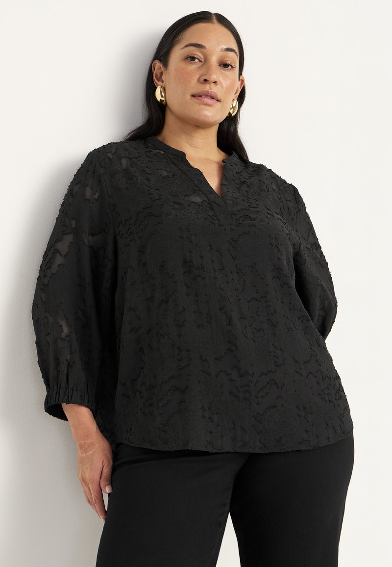 Sort blouse med et struktureret design, V-hals og trekvartærmer. Løs pasform med samlede manchetter; parret med sorte bukser.