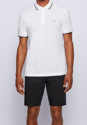Man draagt een wit poloshirt met korte mouwen met zwarte bies en zwarte knielange shorts, staand tegen een effen lichte achtergrond.