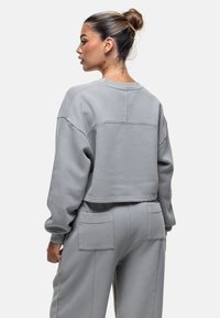 Felpa grigia tagliata con maniche lunghe, realizzata in materiale morbido. Presenta un colletto rotondo e cuciture visibili. La vista posteriore mostra dettagli sulla tasca.