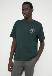 Tmavě zelené bavlněné tričko s kulatým výstřihem, krátkými rukávy a bílou grafikou na levé hrudi, na které je nápis "Cortado Coffee" a hrníček s kávou.