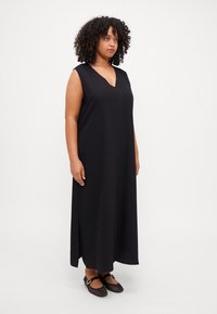 Black sleeveless maxi dress with a V-neckline and side slits. Soft material with a smooth texture. Paired with black shoes featuring a buckle detail.