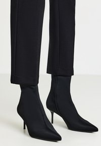 Stivaletti neri aderenti alla caviglia con punte affusolate e sottili tacchi alti indossati con pantaloni neri a tre quarti su uno sfondo chiaro.