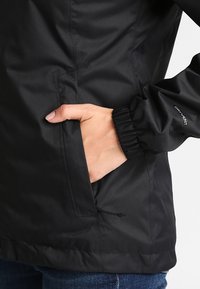 Svart jacka i texturerat tyg med elastiska ärmslut, utrustad med sidoficka och hackad fåll. "DRY VENT"-märke synligt på ärmen.