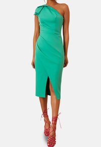 Robe asymétrique à une épaule en vert avec un design drapé et une fente haute sur la cuisse, assortie à des talons roses à lanières. Texture du tissu douce.