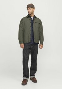 Giubbotto imbottito verde oliva con tasca sul petto, indossato sopra una camicia in denim scuro, abbinato a jeans scuri e stivali stringati marroni.