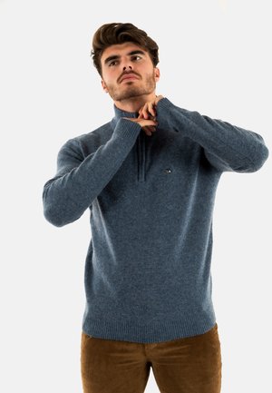 Pull en tricot bleu clair avec col zippé et poignets côtelés. Présente un petit logo sur la poitrine et une texture lisse.