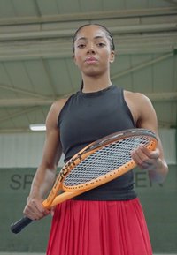 Teniszjátékos egy fekete ütőt tart a kezében, narancssárga kerettel, testhez simuló fekete felsőt és lebegő piros szoknyát visel. Fedett sportkörnyezet.