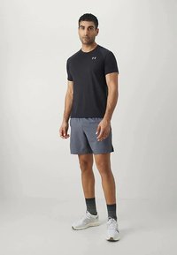 Άντρας που στέκεται με μαύρο αθλητικό μπλουζάκι, γκρι σορτσάκι, μαύρες κάλτσες και άσπρα αθλητικά παπούτσια, μπροστά σε ουδέτερο φόντο.