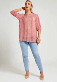 Blusa trasparente rosa chiaro con pois, maniche 3/4, bottone frontale e dettagli arricciati. Abbinata a jeans attillati azzurri.