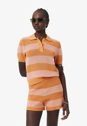 Vrouw die een oranje en roze gestreepte gebreide poloshirt en bijpassende shorts draagt met een zonnebril, staand met een arm over de borst gekruist.