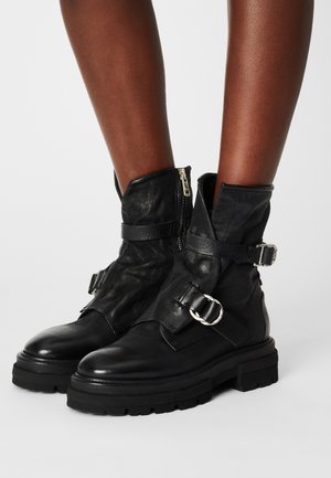 Botas negras de cuero hasta la mitad de la pantorrilla con suelas gruesas, cremalleras laterales y hebillas decorativas, usadas en los pies sobre un fondo liso.