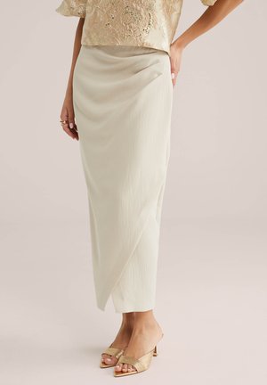 Donna che indossa una gonna midi avvolgente color crema, tacchi aperti dorati con texture, e una blusa in broccato floreale dorato, in piedi con una mano sul fianco.