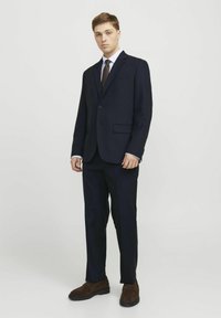 Tume sinine ülikond ühe nupuga sulgemise, sisselõigatud krae, valge särk, pruun lips ja pruunad sametkingad. Kitsas lõige ja siledate fronttsoonidega püksid.