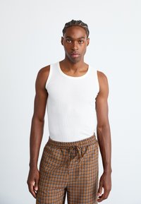 Weißes Baumwoll-Tanktop, kombiniert mit braunen Karo-Hosen mit blauen Akzenten. Das Tanktop hat eine gerippte Textur und ein schlichtes, klassisches Design.
