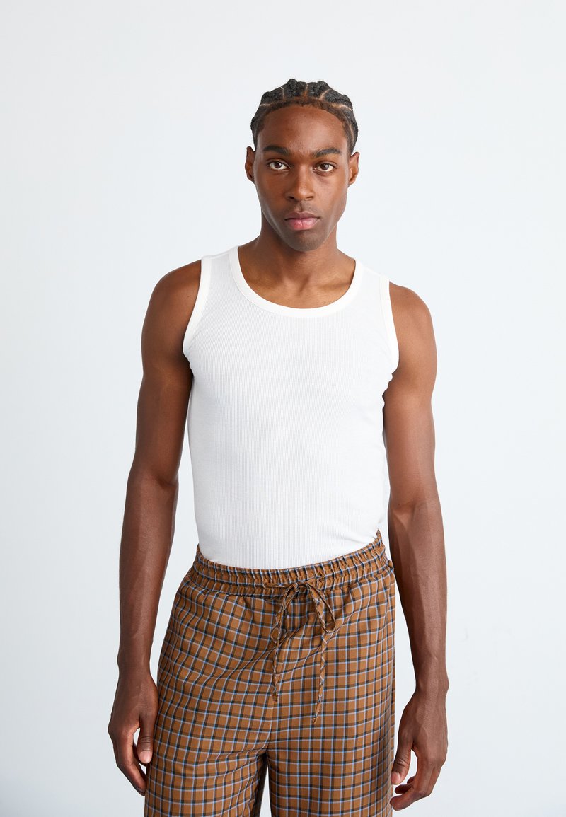 Weißes Baumwoll-Tanktop, kombiniert mit braunen Karo-Hosen mit blauen Akzenten. Das Tanktop hat eine gerippte Textur und ein schlichtes, klassisches Design.