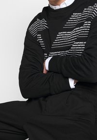 Svart sweatshirt med vit text som detalj, "Emporio Armani", i olika storlekar och orienteringar, med rund hals och långa ärmar.