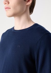 Pulover tricotat bleu marin cu guler rotund și un mic logo CK brodat pe piept. Textură netedă, formă ajustată și umeri fără cusături.