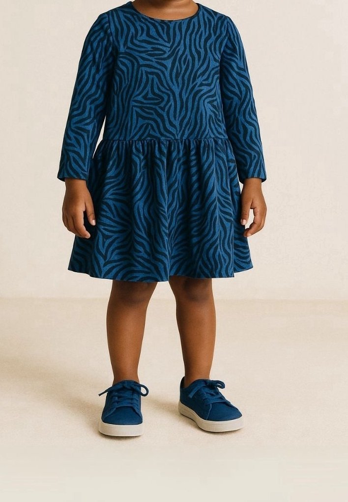 Barn klädd i en långärmad blå klänning med mörka zebraränder och matchande blå sneakers med vita sulor, stående på ett beige golv.