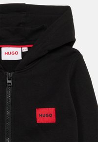 Μαύρο φούτερ με φερμουάρ μπροστά, κόκκινο τετράγωνο πατς στο στήθος με την ένδειξη "HUGO" και εσωτερική ετικέτα στον λαιμό που δείχνει το όνομα της μάρκας και το μέγεθος.