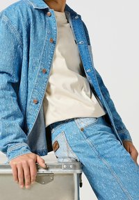 Giacca di jeans e jeans in azzurro chiaro con una finitura usurata. Presenta bottoni di rame, una camicia beige sotto e una toppa in pelle beige sui jeans.