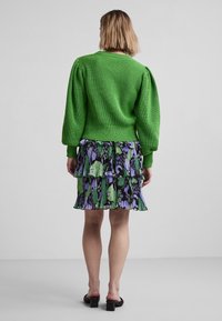 Pull vert tricoté avec manches bouffantes associé à une robe à volants multicouches ornée d'un motif floral et de feuilles en vert, noir et violet.
