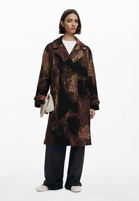 Přehnaně velký kabát s tmavým vzorem v odstínech hnědé a černé, s širokým límcem, dlouhými rukávy a páskem v pase.