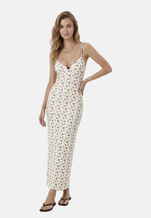 Donna con un abito lungo bianco a fiori senza maniche con sottili spalline, indossa una collana dorata a catena e infradito marroni, in piedi con una mano sul fianco.