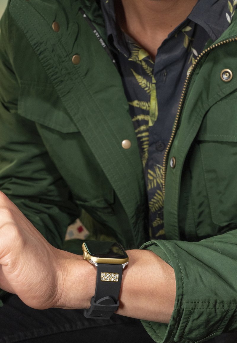 Smartwatch with a black rubber strap and a gold-tone metal case; square display; worn by a person in a green jacket and patterned shirt.