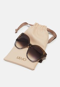 Overdimensjonerte brune solbriller med gradientlinser, som hviler på en beige pose med snøring og gull "LIU•JO"-logo.