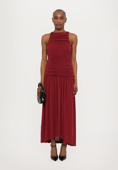 Burgunderfarbene, ärmellose Maxikleid mit gerüschtem Oberteil und fließendem Rock, ergänzt durch eine kleine schwarze Handtasche und schwarze spitze Pumps.