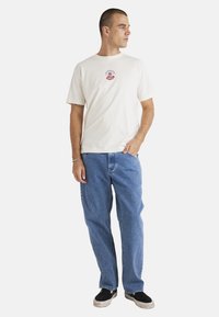 Camiseta de algodón blanca con un diseño gráfico en rojo y azul en el pecho, combinada con jeans de mezclilla azul claro. Modelo con zapatos negros tipo slip-on.