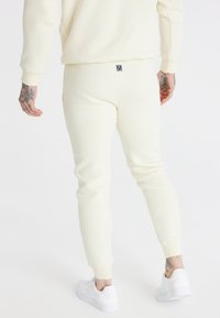 Krämfärgad joggerbyxa i mjukt tyg med avsmalnande ben, ribbade muddar och en liten logotagg på baksidan, tillsammans med vita sneakers.