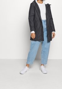 Svart vattentät jacka med huva, framknäppning med tryckknappar och sidofickor; bärs över en vit ribbad topp och ljusblå baggy jeans.