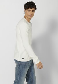 Witte, ribgebreide lange mouwen shirt met een ronde halslijn, gecombineerd met blauwe jeans. Klein merklabel zichtbaar aan de zoom.