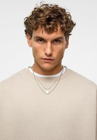 Beige sweatshirt met een ronde hals, gelaagd over een witte shirt. Twee zilveren kettingen met een klein rechthoekig hanger hangen om de nek.