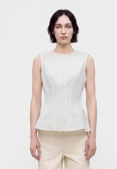 Jeune personne aux cheveux courts et foncés portant un haut blanc ajusté sans manches et un pantalon beige clair sur un fond uni clair.