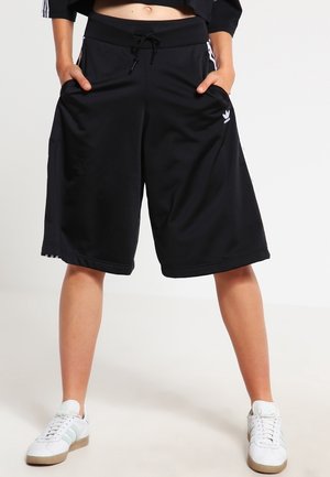 Persona que lleva pantalones cortos negros de Adidas hasta la rodilla con rayas blancas laterales y zapatillas blancas, de pie con las manos en los bolsillos.