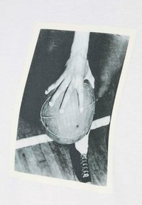 T-shirt bianco con una grafica in bianco e nero di una mano che afferra un pallone da basket. L'immagine è incorniciata da un bordo crema.
